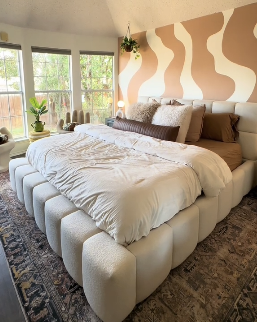 A bedroom with a large bed with pillows, patterned wall, ceiling fan, and windows offering natural light