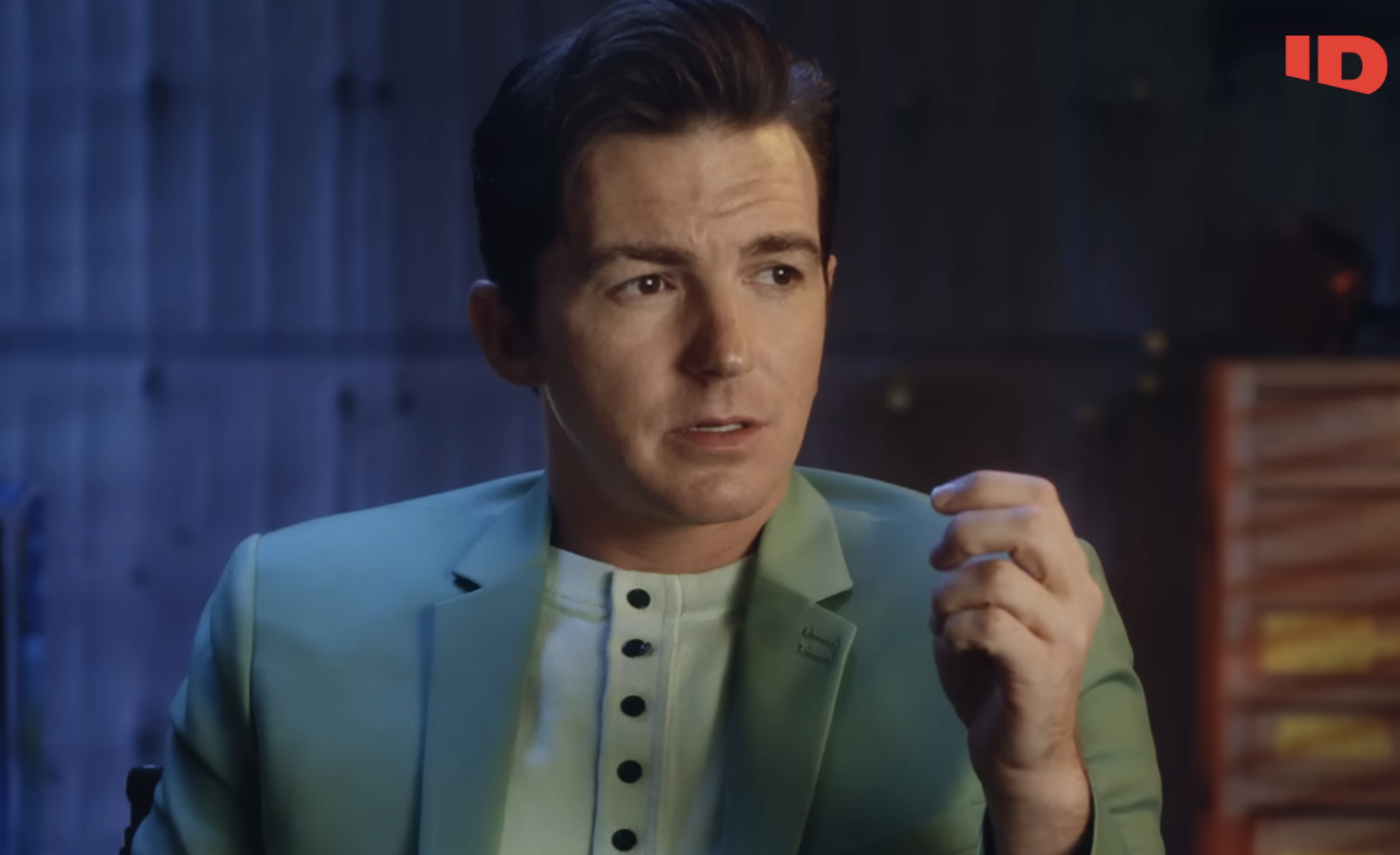 Drake Bell sits for an interview, wearing a green jacket and white shirt, with a microphone clipped on