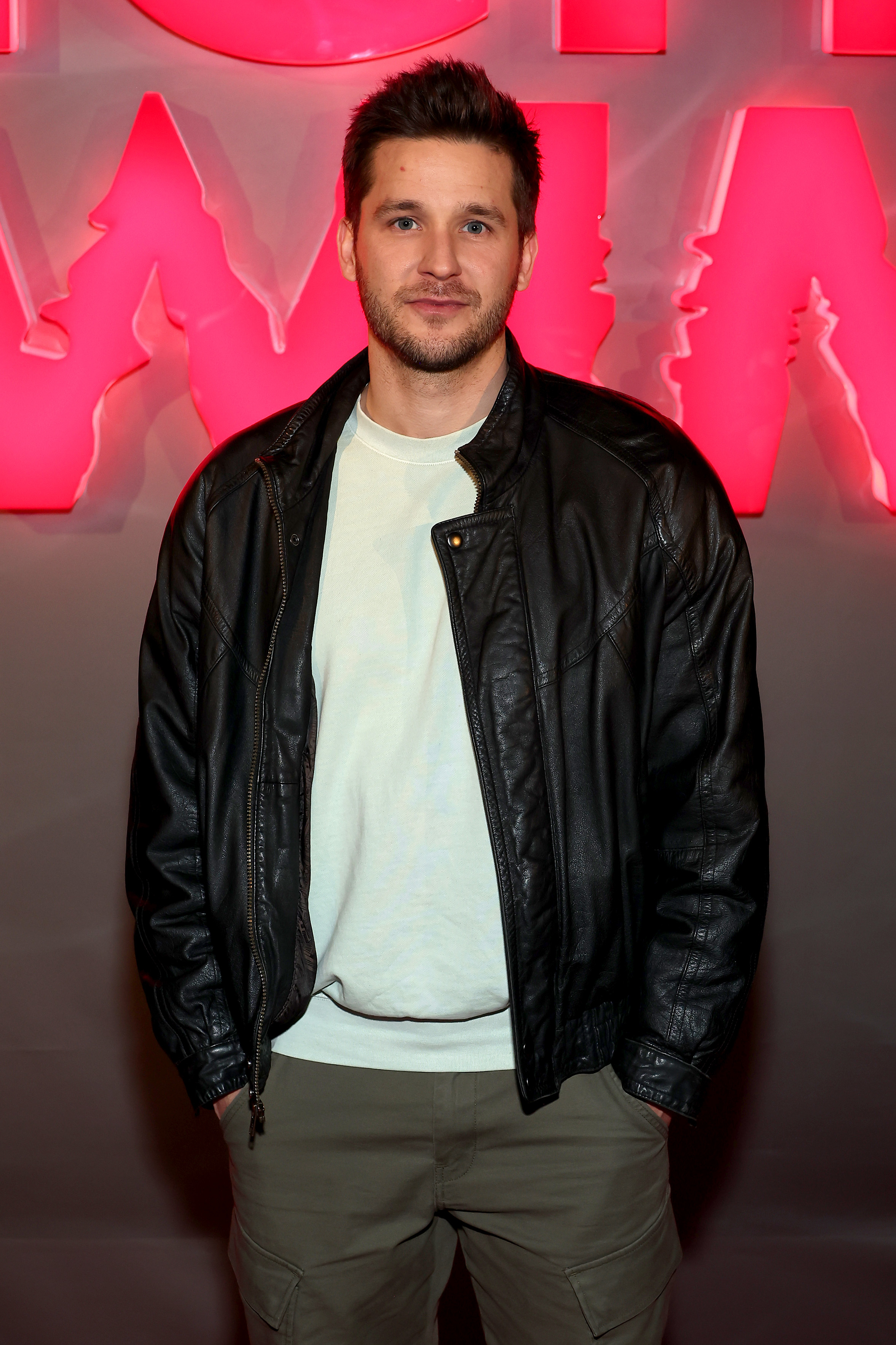 A closeup of adult Devon posing on the red carpet in a leather jacket, tee, and slacks