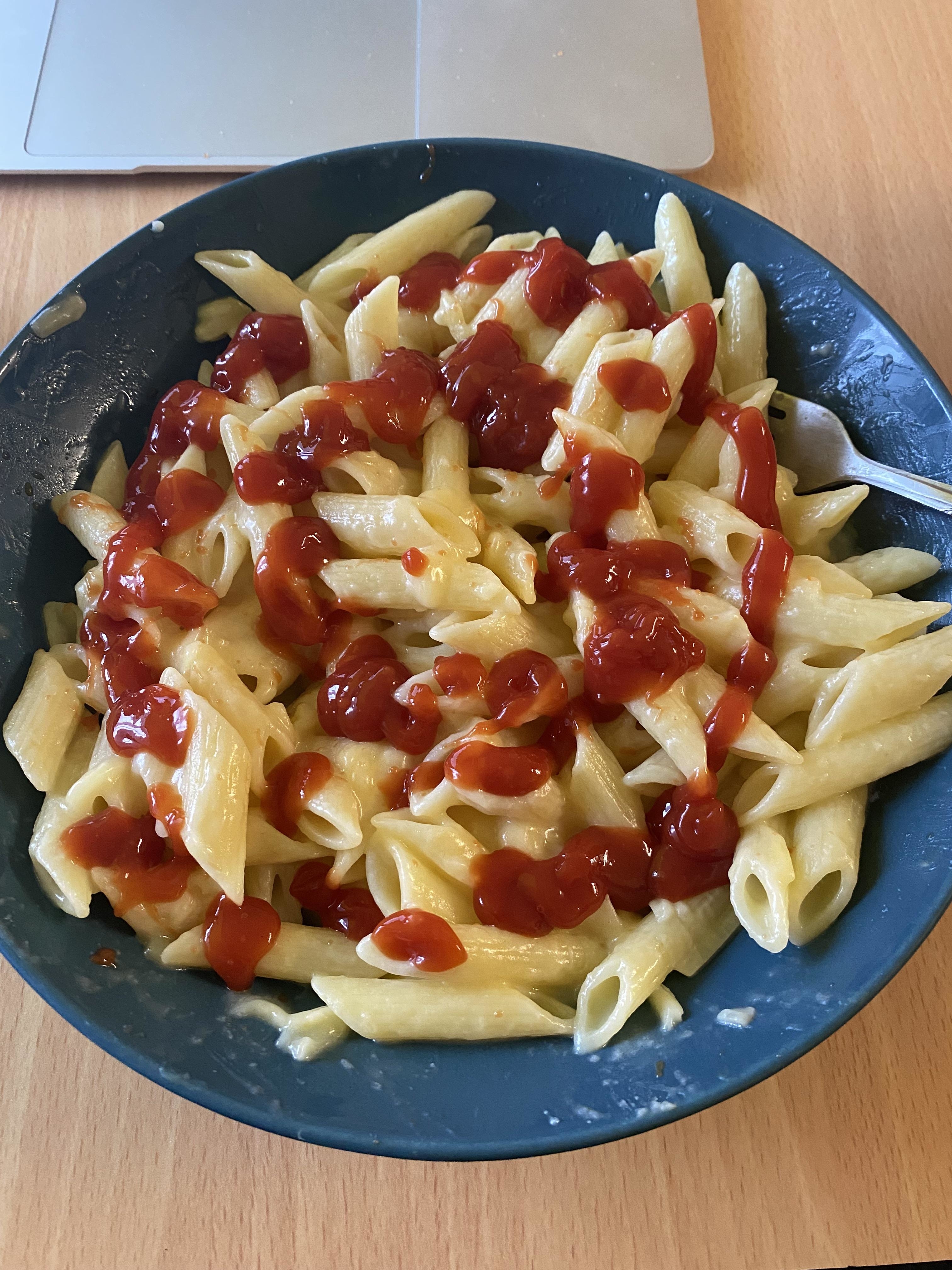 A plate of penne pasta with dollops of ketchup on a wooden table, next to a laptop