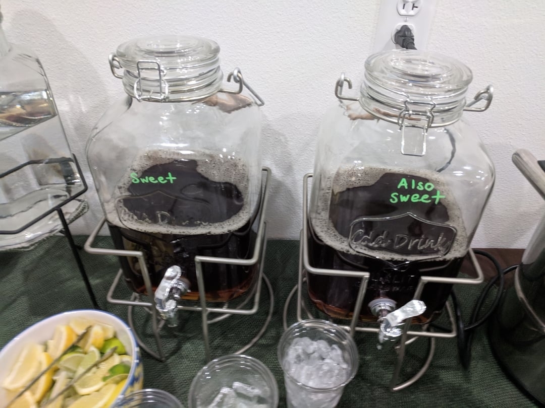 Two beverage dispensers labeled &quot;Sweet&quot; and &quot;Also Sweet&quot; with ice cubes and cups in front