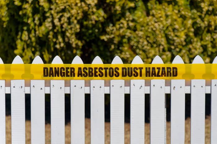 Yellow caution tape with &quot;DANGER ASBESTOS DUST HAZARD&quot; across a white picket fence, warning of hazardous material