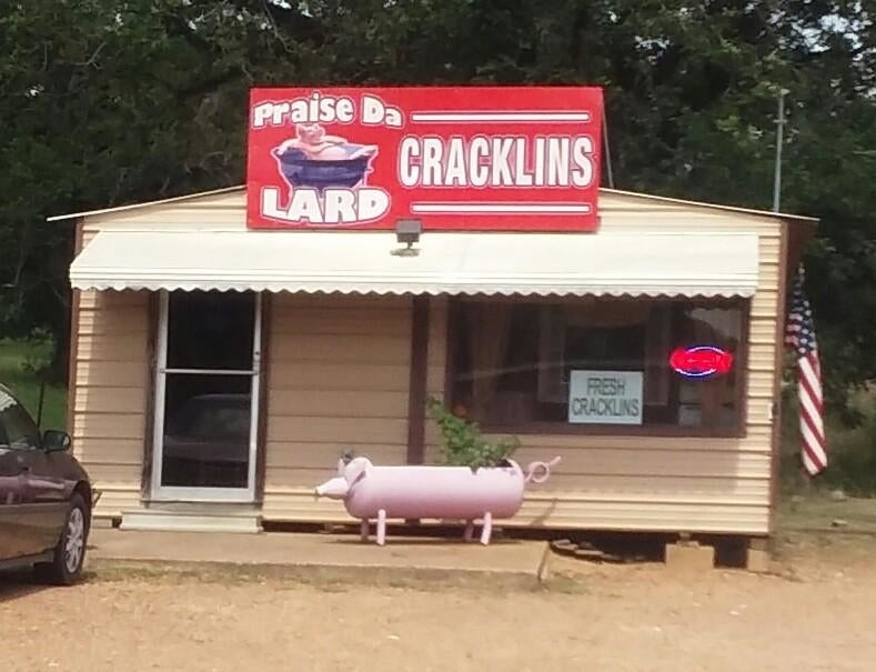 A small building with a sign that reads &quot;praise Da LARD CRACKLINS&quot; and features a pig-shaped bench in the front