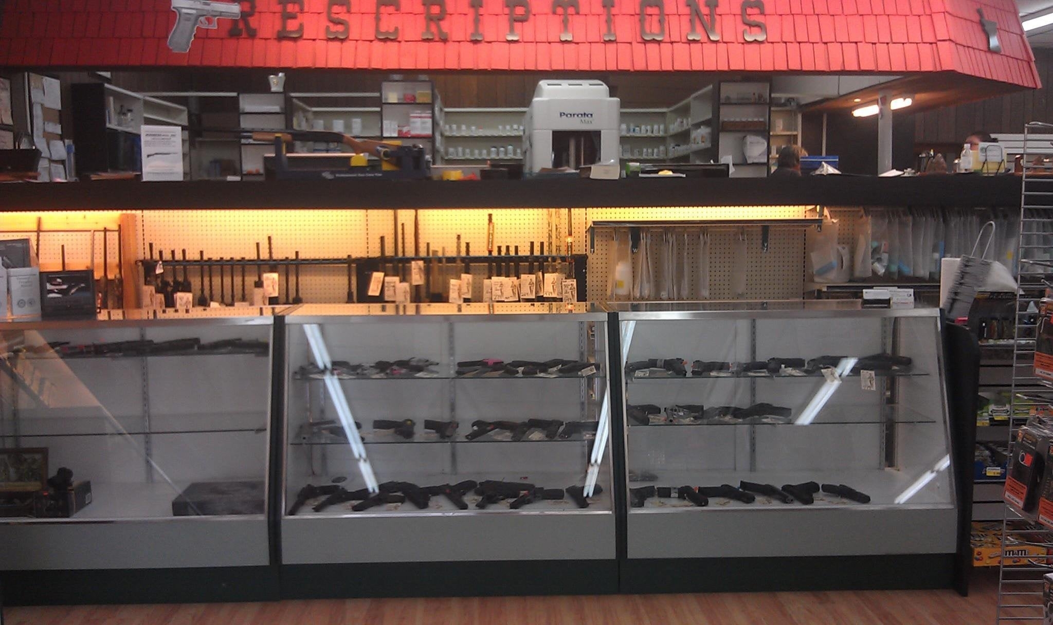 Gun counter with firearms displayed under glass, adjacent to a prescriptions sign in a store