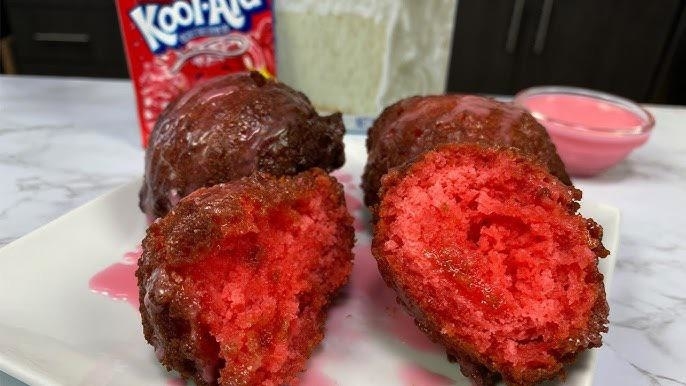 Three deep-fried Kool-Aid balls on a plate with a packet of Kool-Aid and a cup of sauce in the background