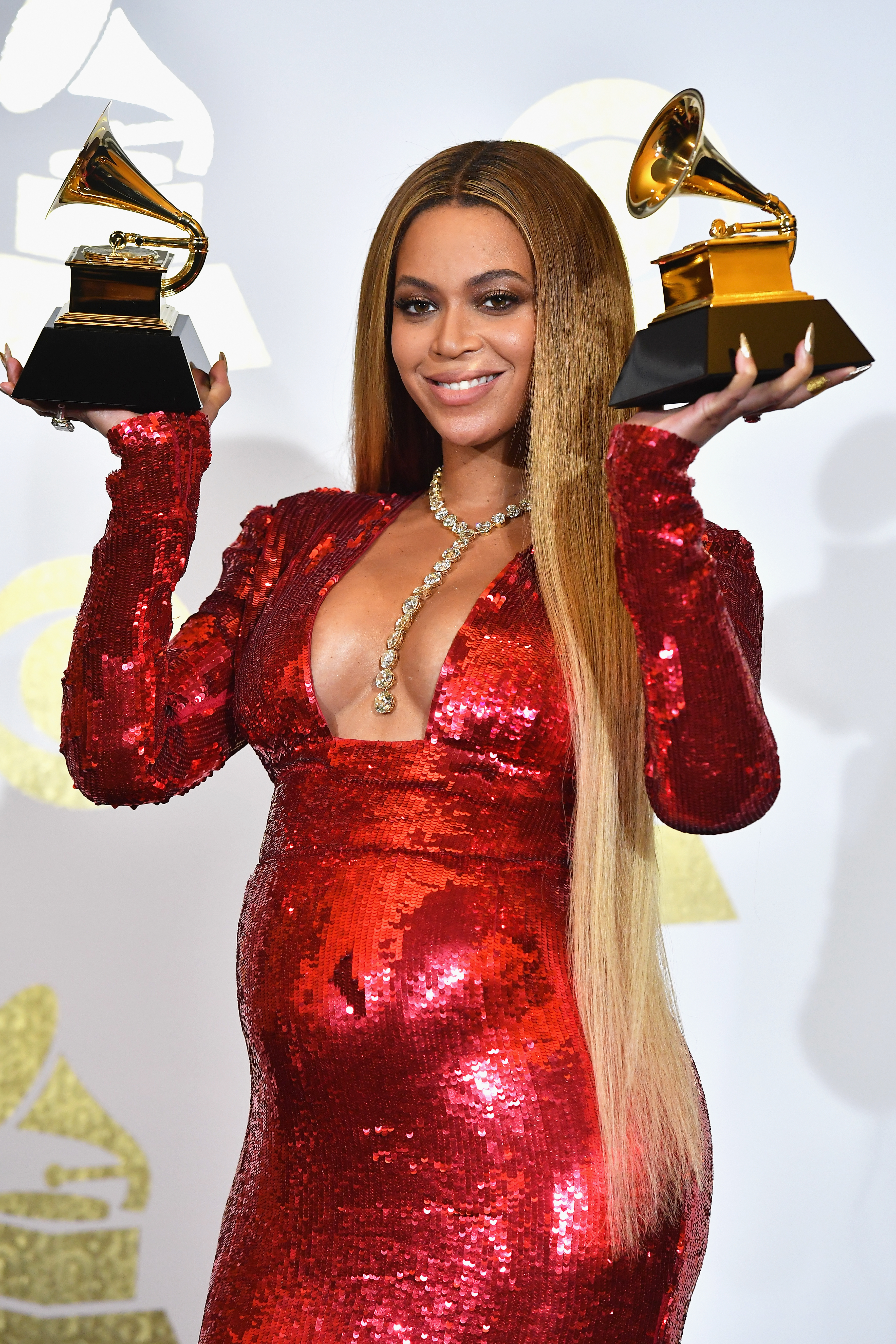 Beyoncé smiling, holding Grammy Awards, wearing a sequined dress with a v-neckline and long, straight hair