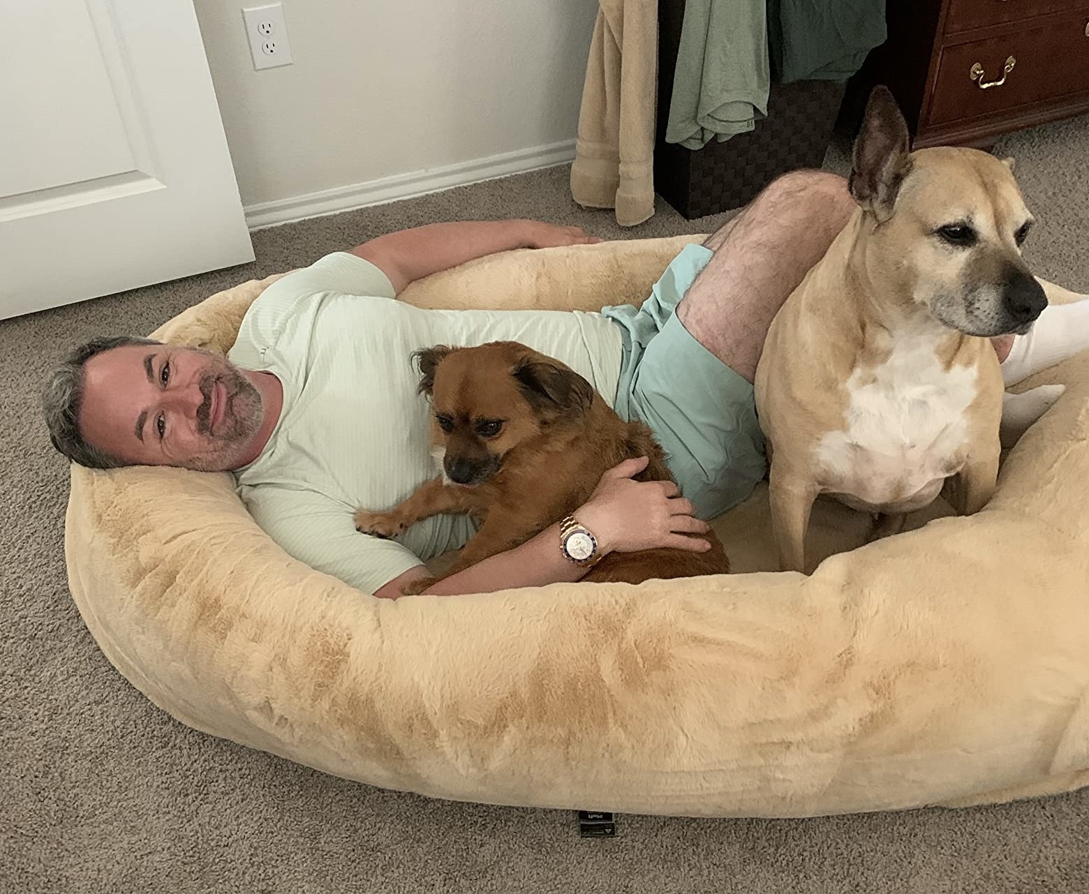 person lying in a large pet bed with two dogs, looking happy and relaxed