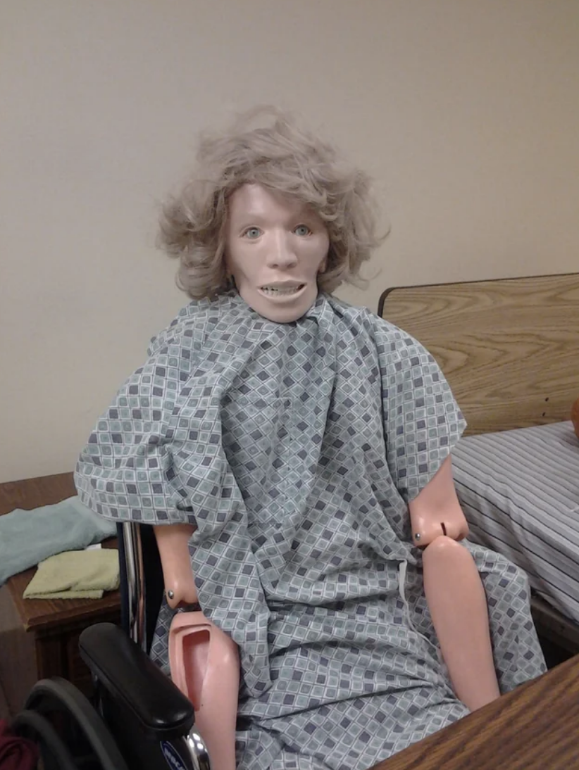 Mannequin in a wheelchair wearing a hospital gown, positioned indoors for training