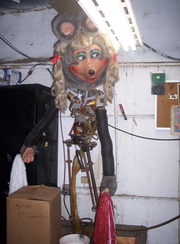 An animatronic figure with a mouse head and beige dress in a workshop setting