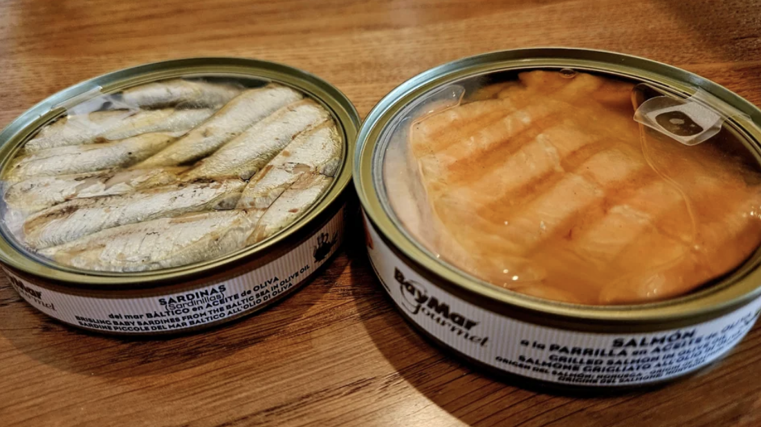 Two cans of seafood, one with sardines and the other with salmon, opened on a wooden surface