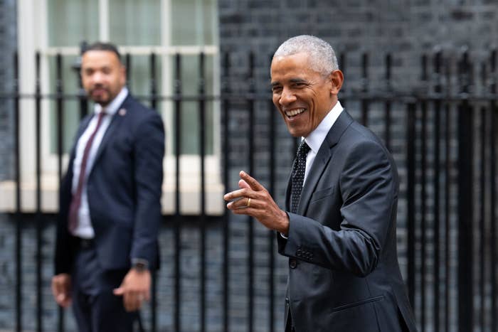 Former President Barack Obama in a suit, smiling and gesturing, with a security agent in the background