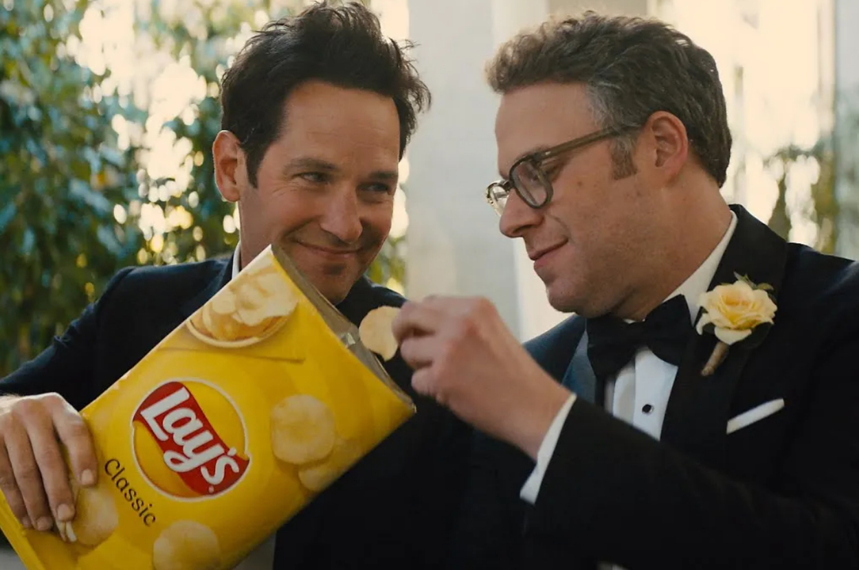 Two men in suits share a bag of Lay's chips, smiling at each other