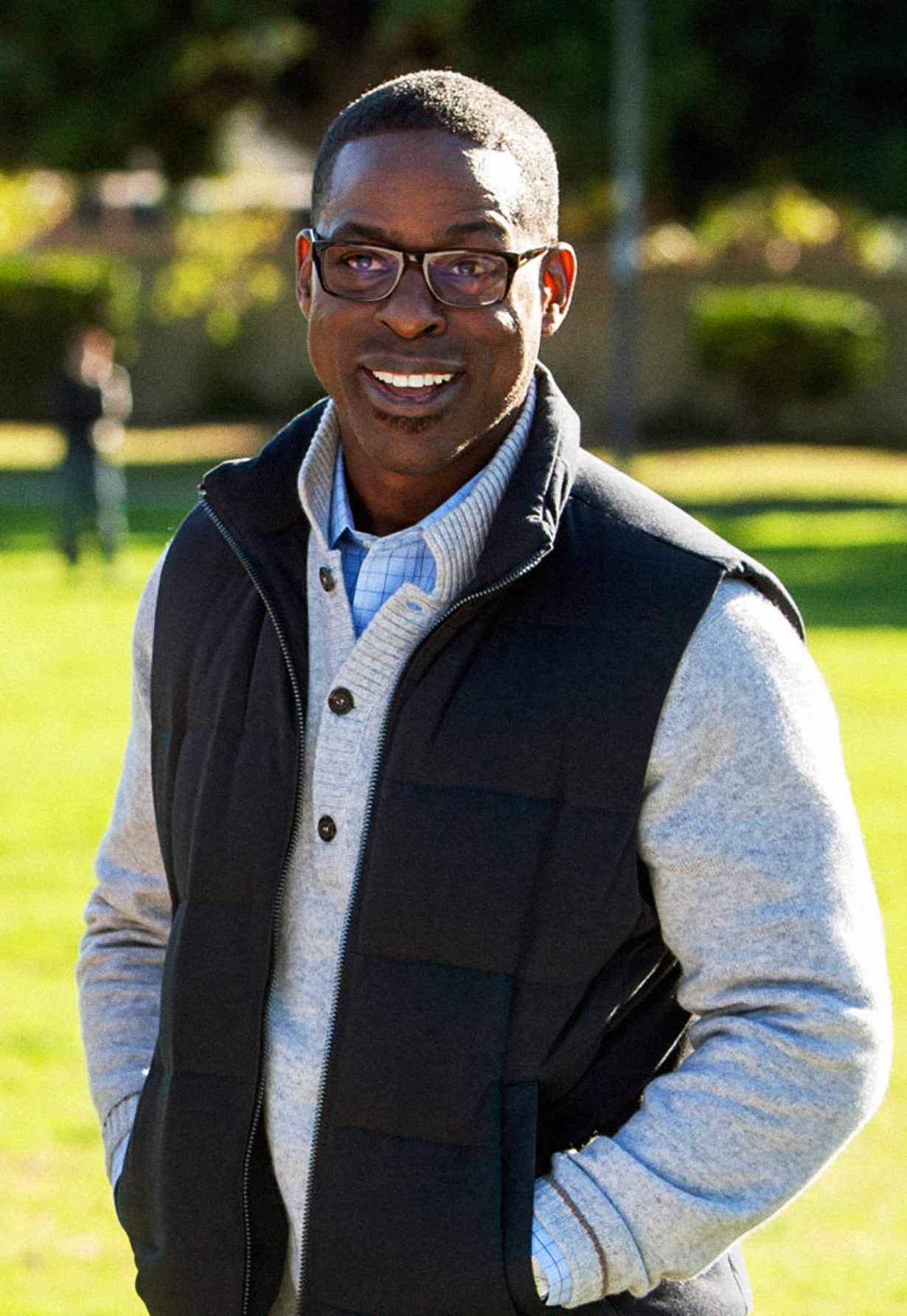 closeup of him wearing glasses and a vest over a sweater as he stands outside