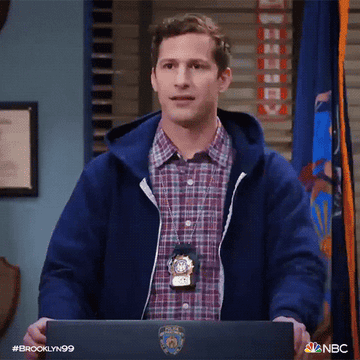 Jake Peralta from Brooklyn Nine-Nine in a police station, looking surprised as he stands up from his desk