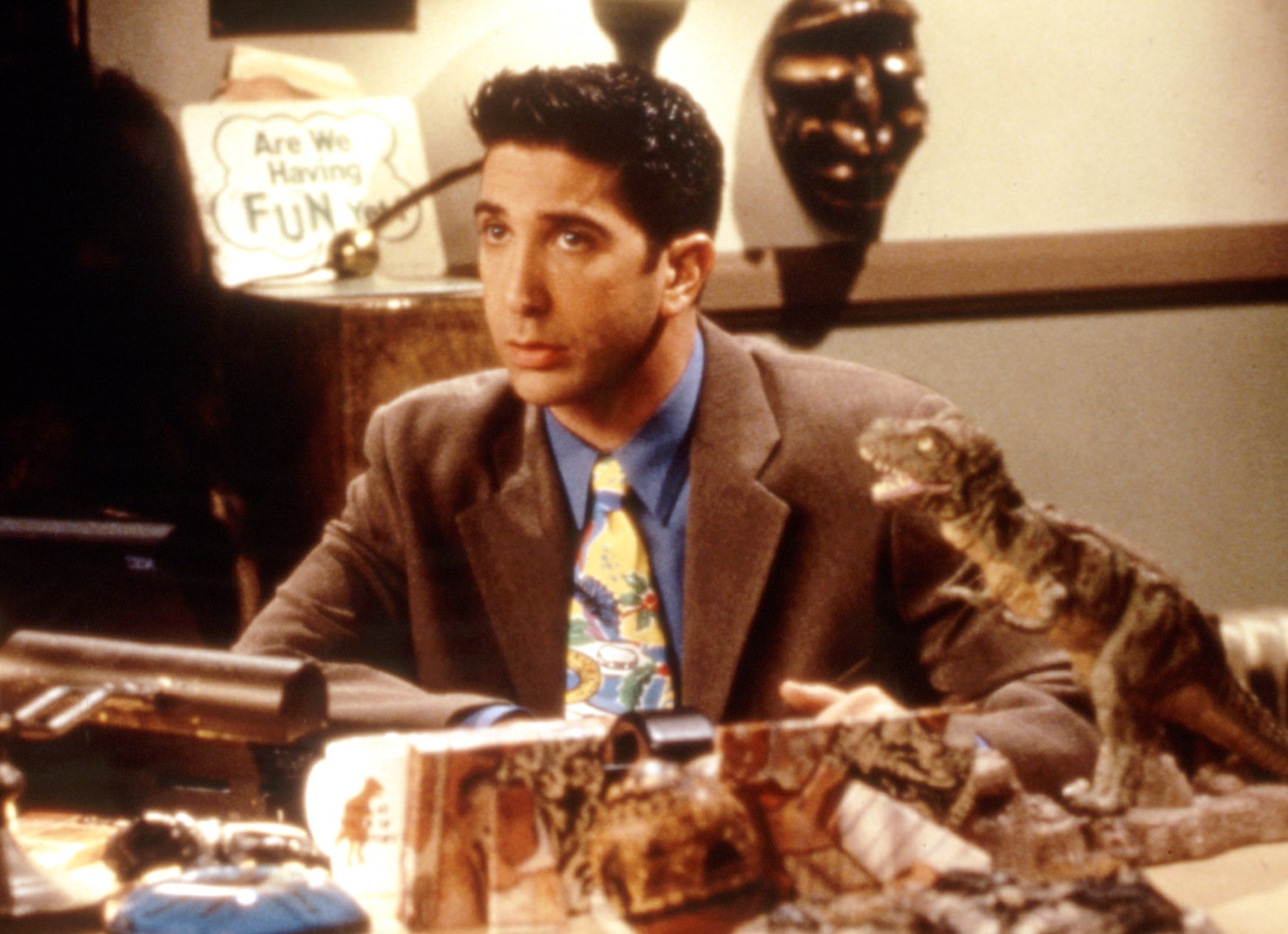 Ross from Friends sits at a desk surrounded by dinosaur artifacts, wearing a jacket and tie with a dinosaur-themed shirt