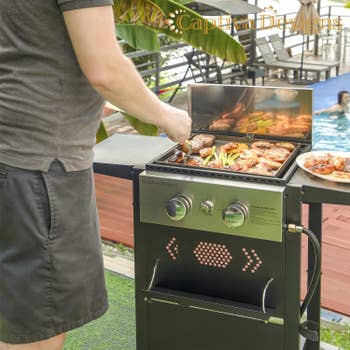 A person is grilling food on an outdoor barbecue with others relaxing in a pool in the background
