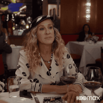 Sarah Jessica Parker as Carrie Bradshaw in a scene, wearing a stylish outfit with a hat, seated at a table