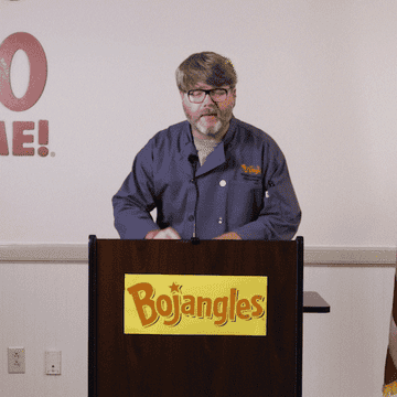 Gif of a person standing at a Bojangles&#x27; podium wearing a casual jacket and glasses saying, &quot;It&#x27;s so cluckin&#x27; good&quot;