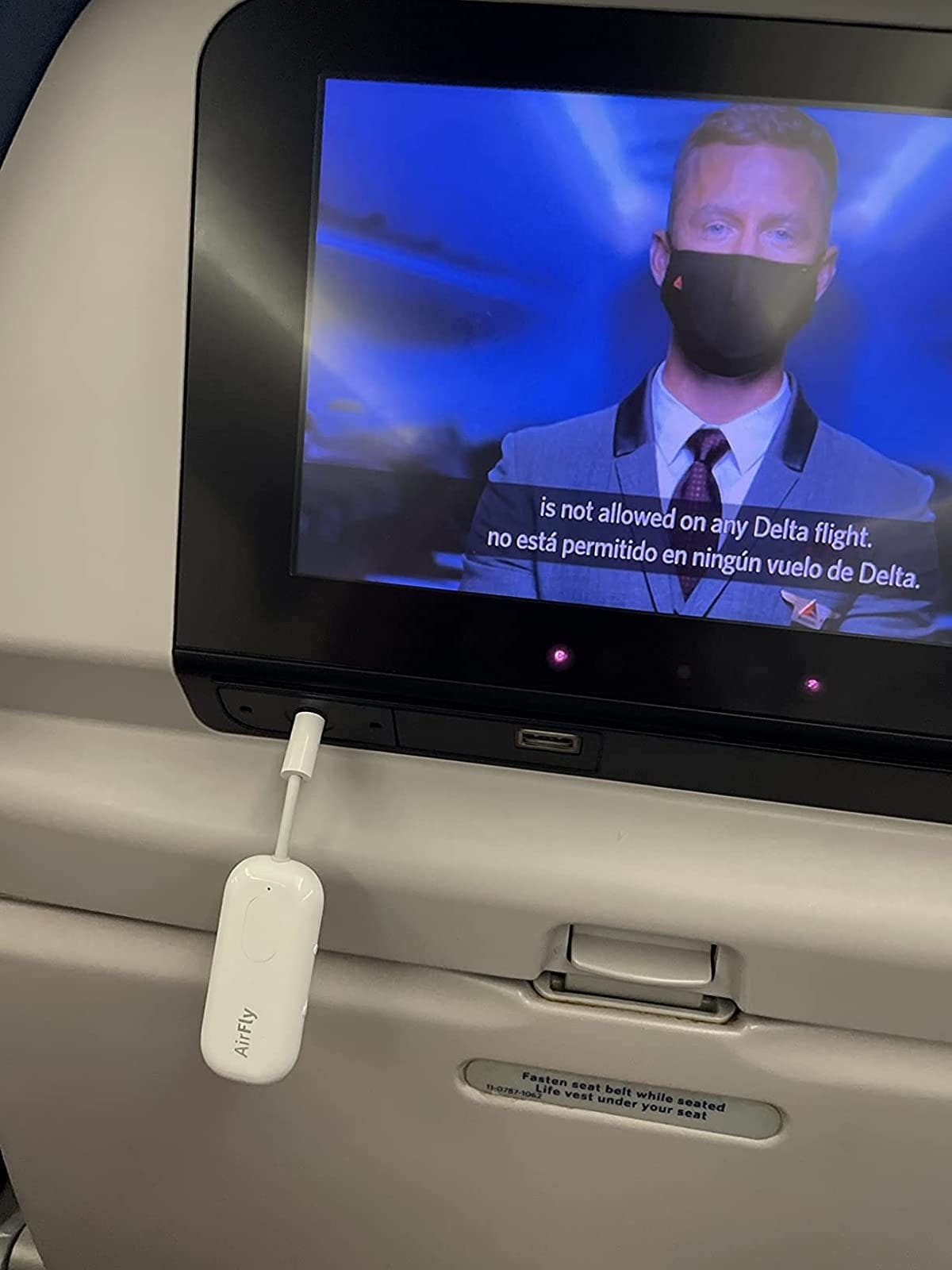 In-flight screen showing safety video with a masked actor, captioned about Delta flight rules, and a charger plugged in below