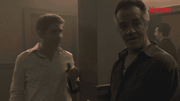 Two men in a scene from "The Sopranos," one holding a beer bottle, conversing in a room