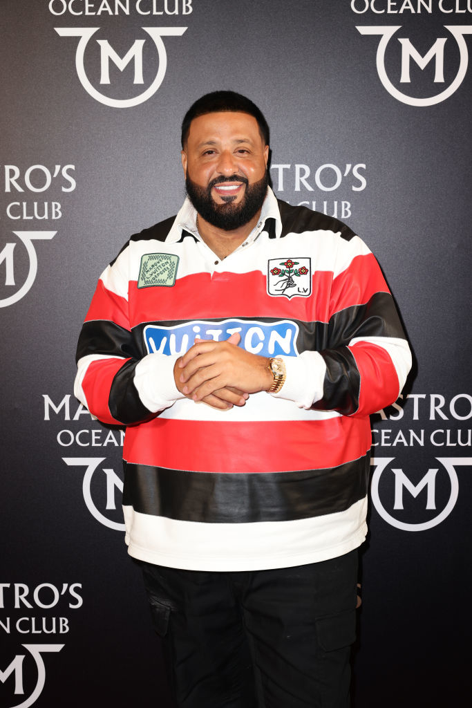 DJ Khaled in a striped shirt and black pants, smiling with hands crossed, at an event