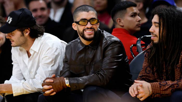 Bad Bunny seated at a basketball game wearing eyeglasses and a leather jacket