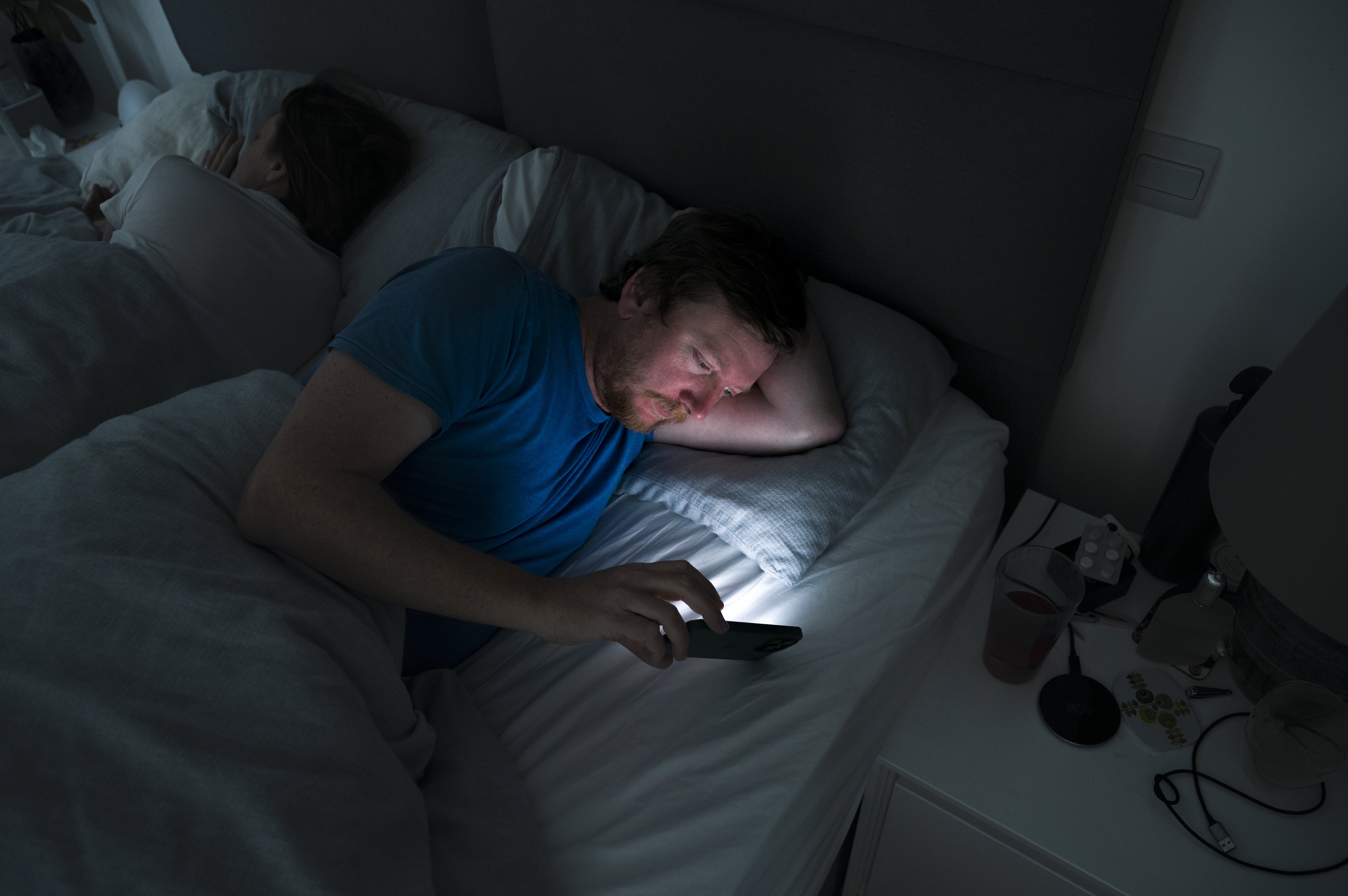 Man in bed using phone with another person asleep beside him, highlighting the impact of screen time on sleep