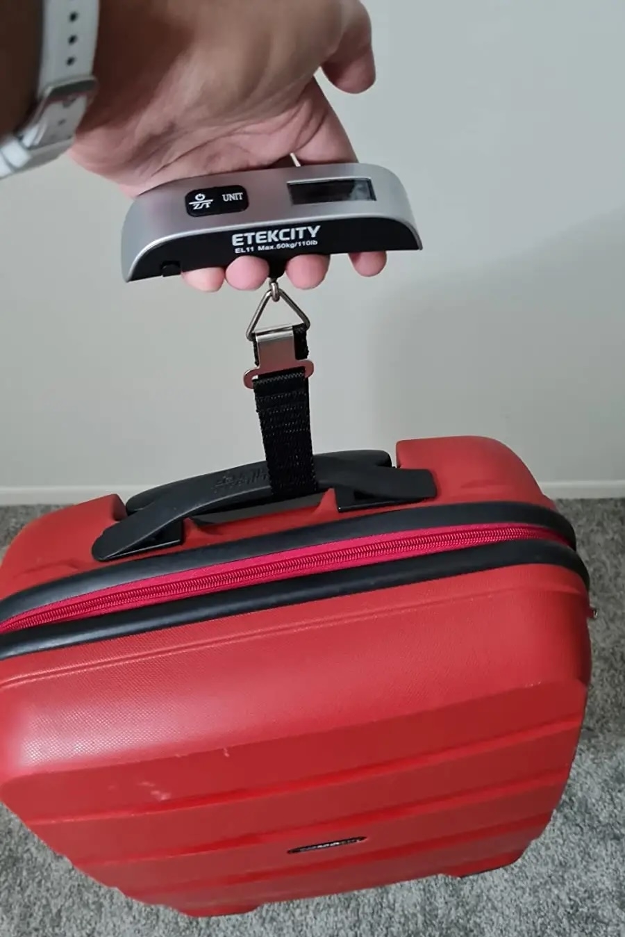Person using a portable digital luggage scale to weigh a red suitcase