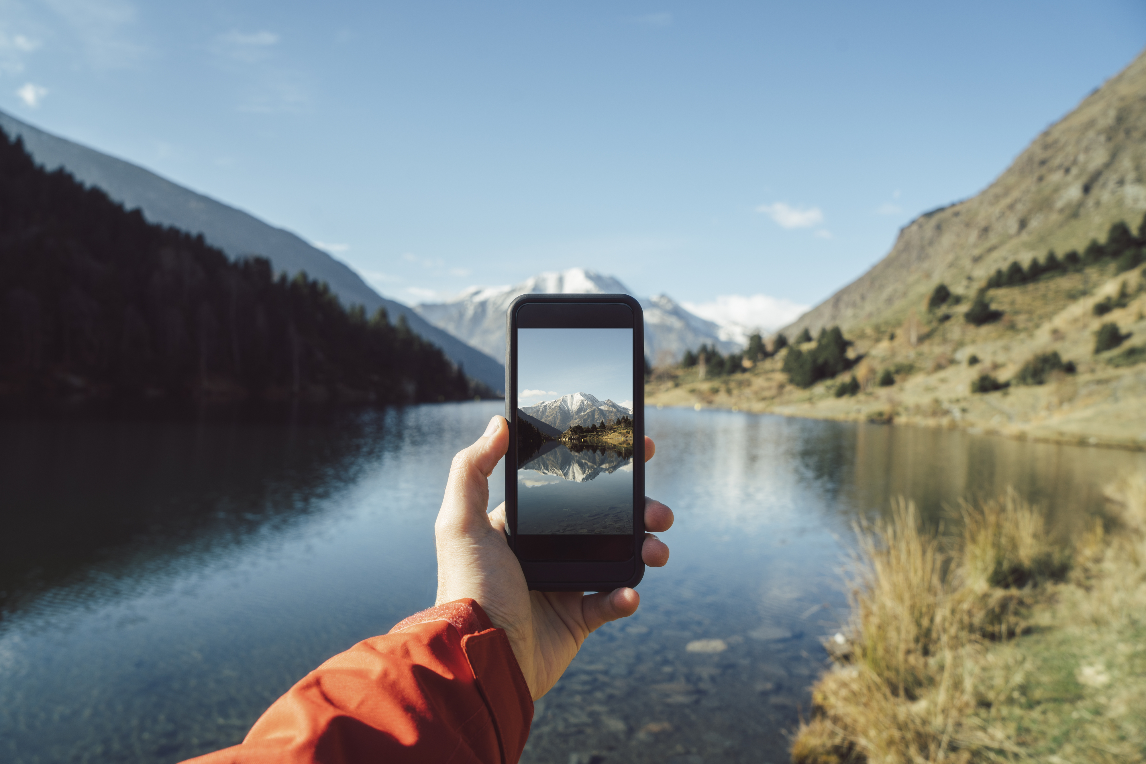 Hand holding a smartphone capturing a mountain and lake scene