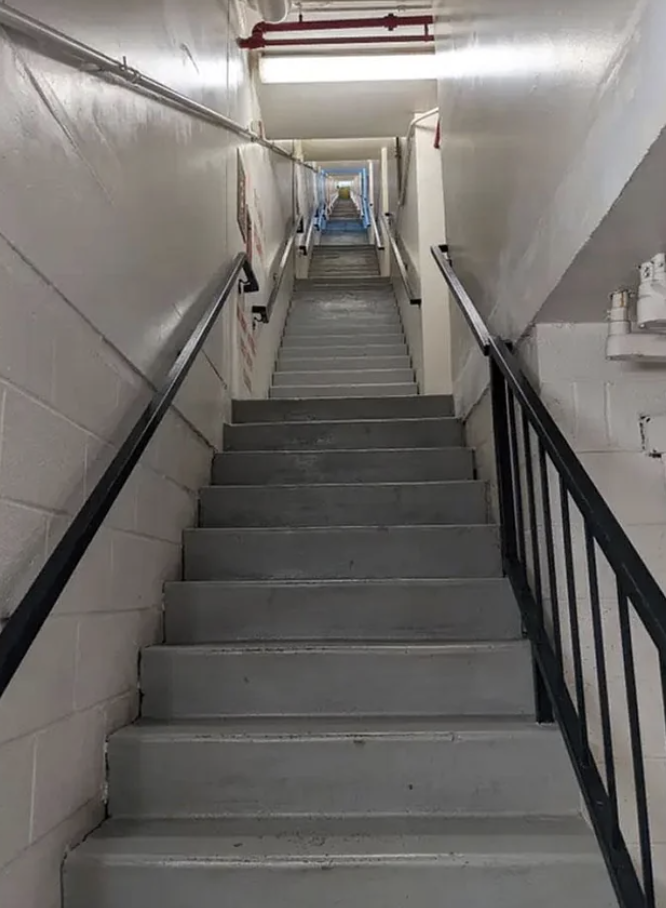 Narrow staircase with steep steps and handrails leading up to a bright exit