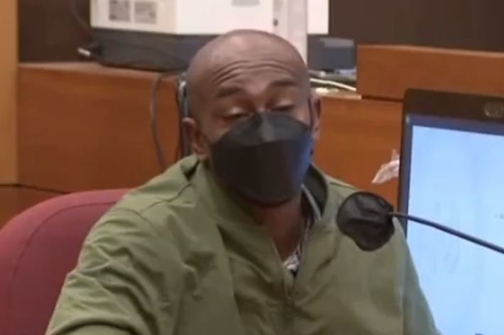 Man with a mask sitting in a courtroom, speaking into a microphone