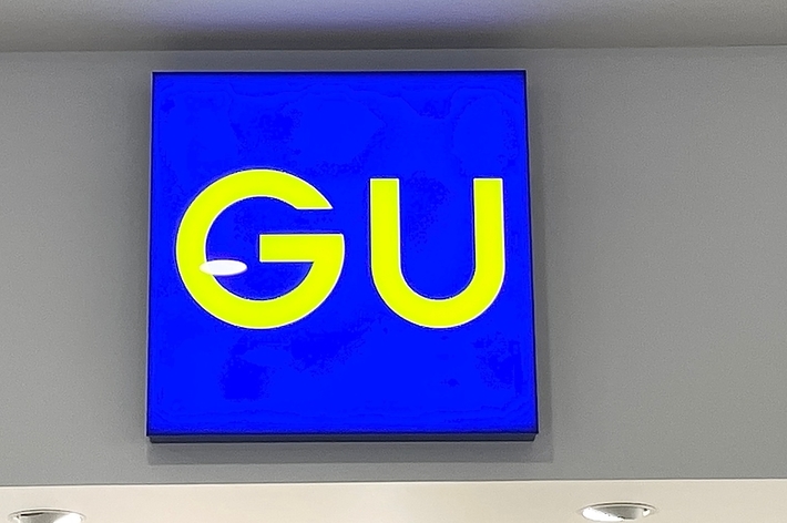 Sign with “GU” lettering on a blue background, mounted on a wall with ceiling fixtures visible