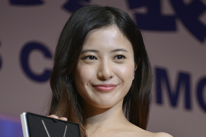 女性が黄色いドレスを着てネックレス入りの箱を持つ手に笑顔で対応している。