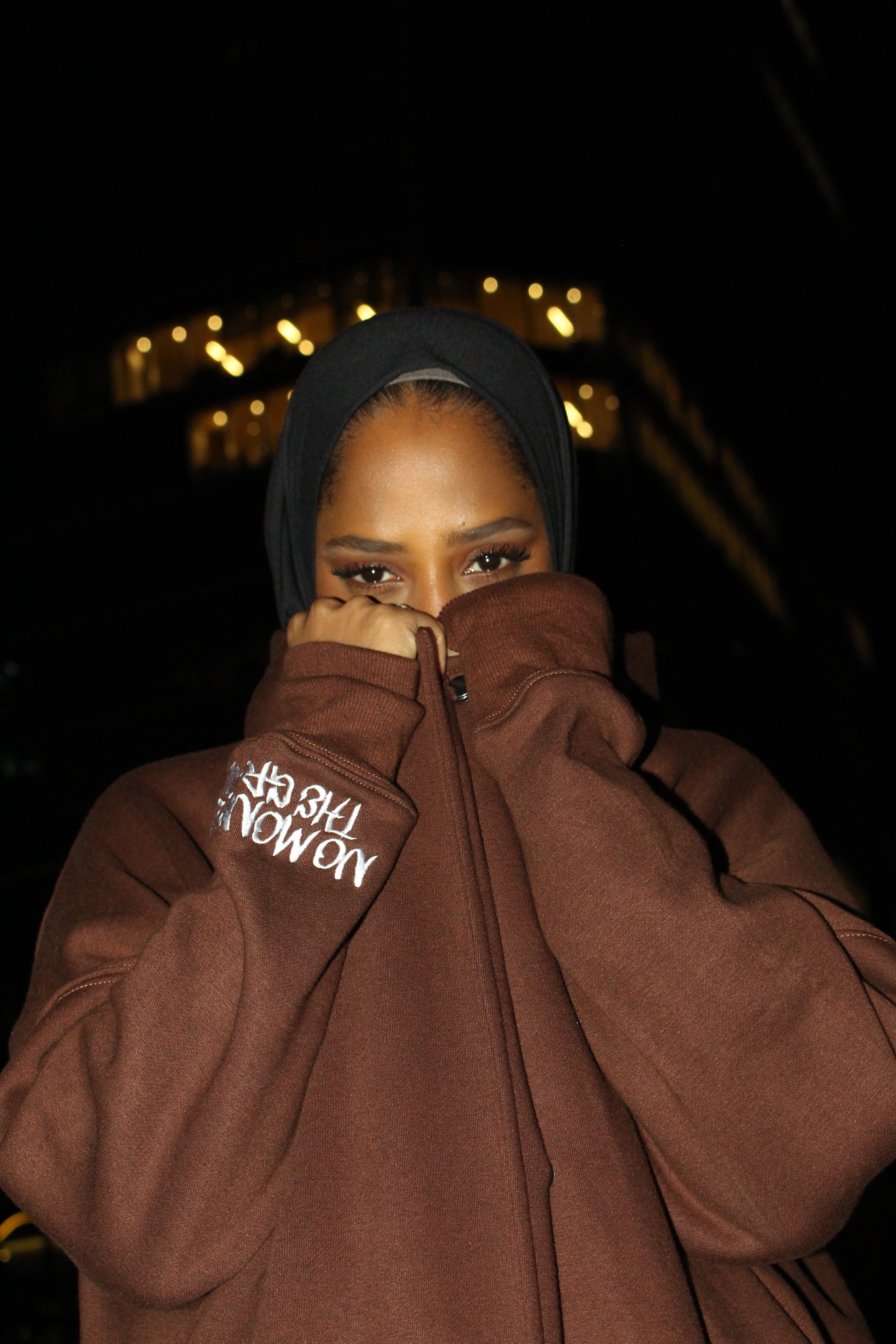 A woman in a hoodie abaya covering lower face with her hands, standing in the night with lights in the background