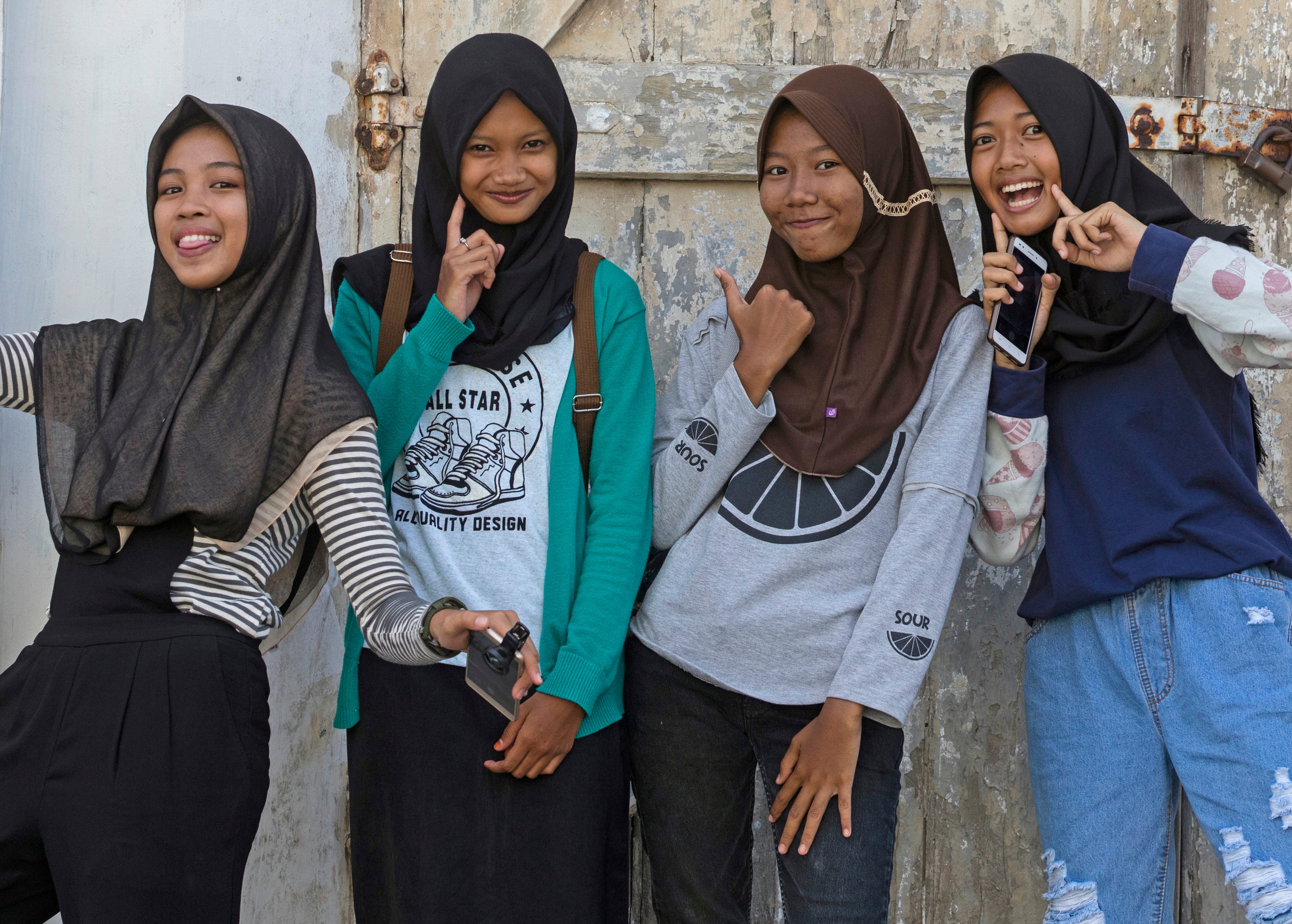 Four girls posing with smiles against a wall, one making a peace sign, others with playful gestures
