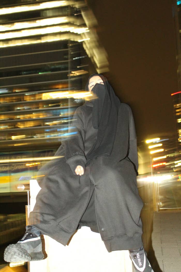 Saeedah sitting at night with lights streaking in the background, wearing a hoodie abaya and niqab
