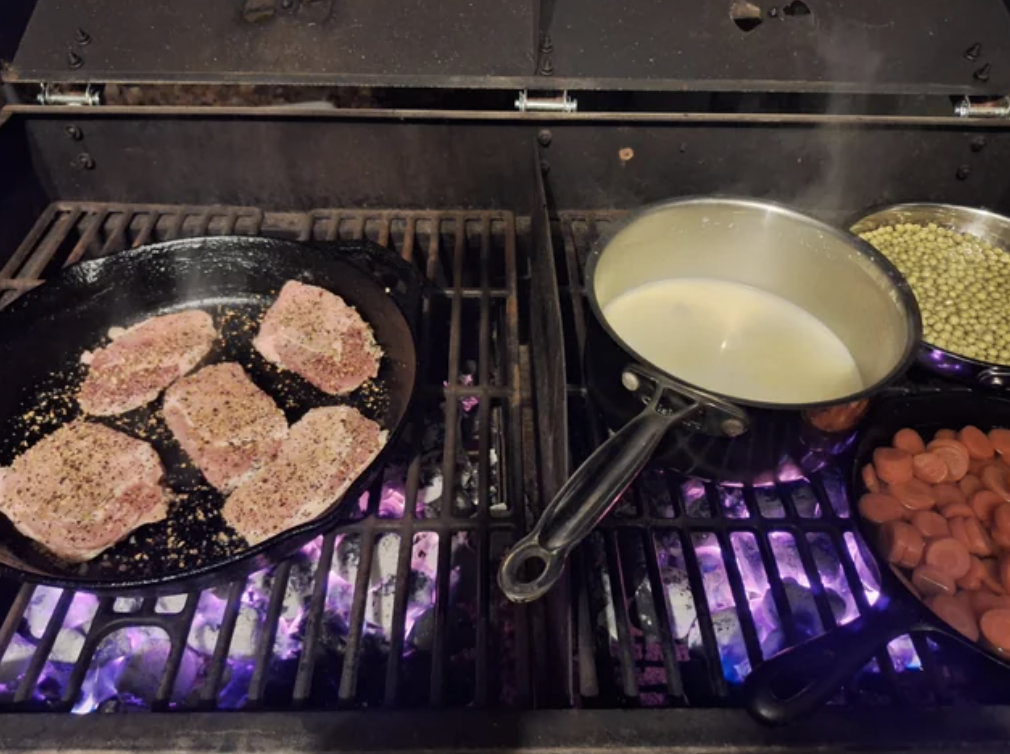 Cooking meal with four seasoned pork chops in a skillet, and pots of milk and peas, hot dogs on a stove