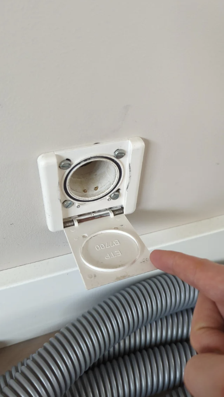 A person&#x27;s finger pointing at an open, empty wall-mounted vacuum inlet, with a coiled hose below it