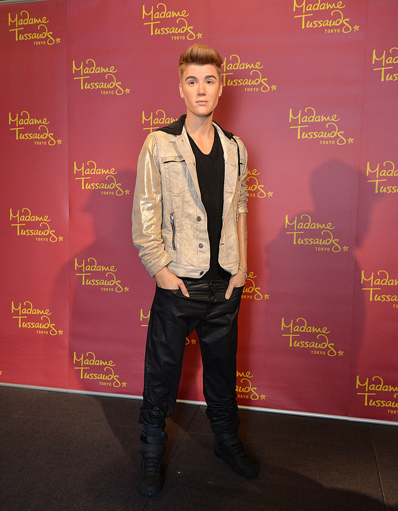 Wax figure of Justin Bieber in a shirt, pants, and a light jacket, standing in front of a branded backdrop
