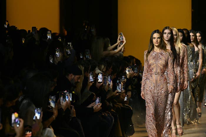 Models walk the runway in sequined dresses at a fashion show with audience taking photos