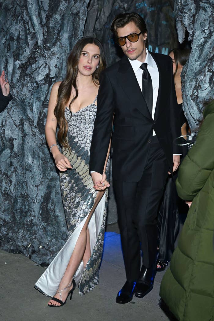 The two walking hand in hand; one in a patterned gown and the other in a suit with sunglasses
