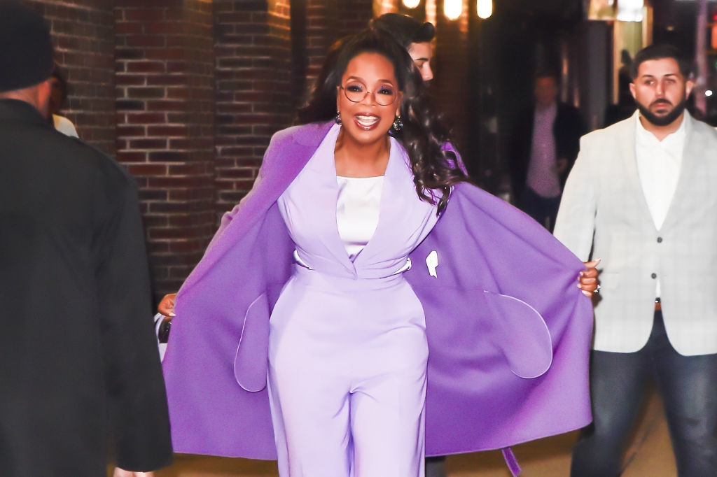 Oprah Winfrey in a stylish suit and matching cape, smiling, walking outdoors