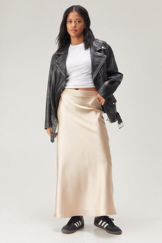 Woman in a leather jacket, white top, and satin skirt paired with sneakers, posing for a shopping article