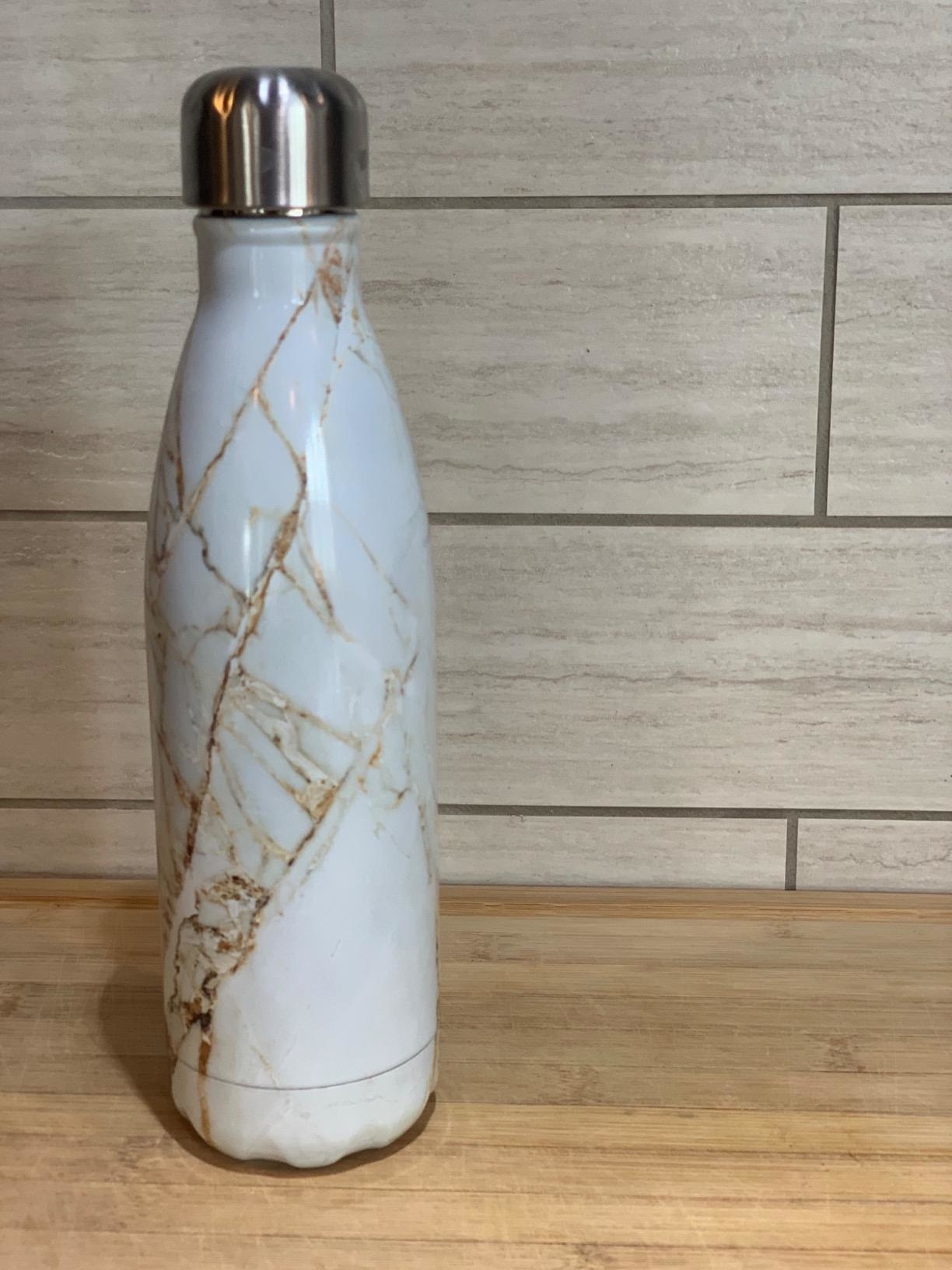 Insulated bottle with a marble design on a wooden surface against a tiled wall