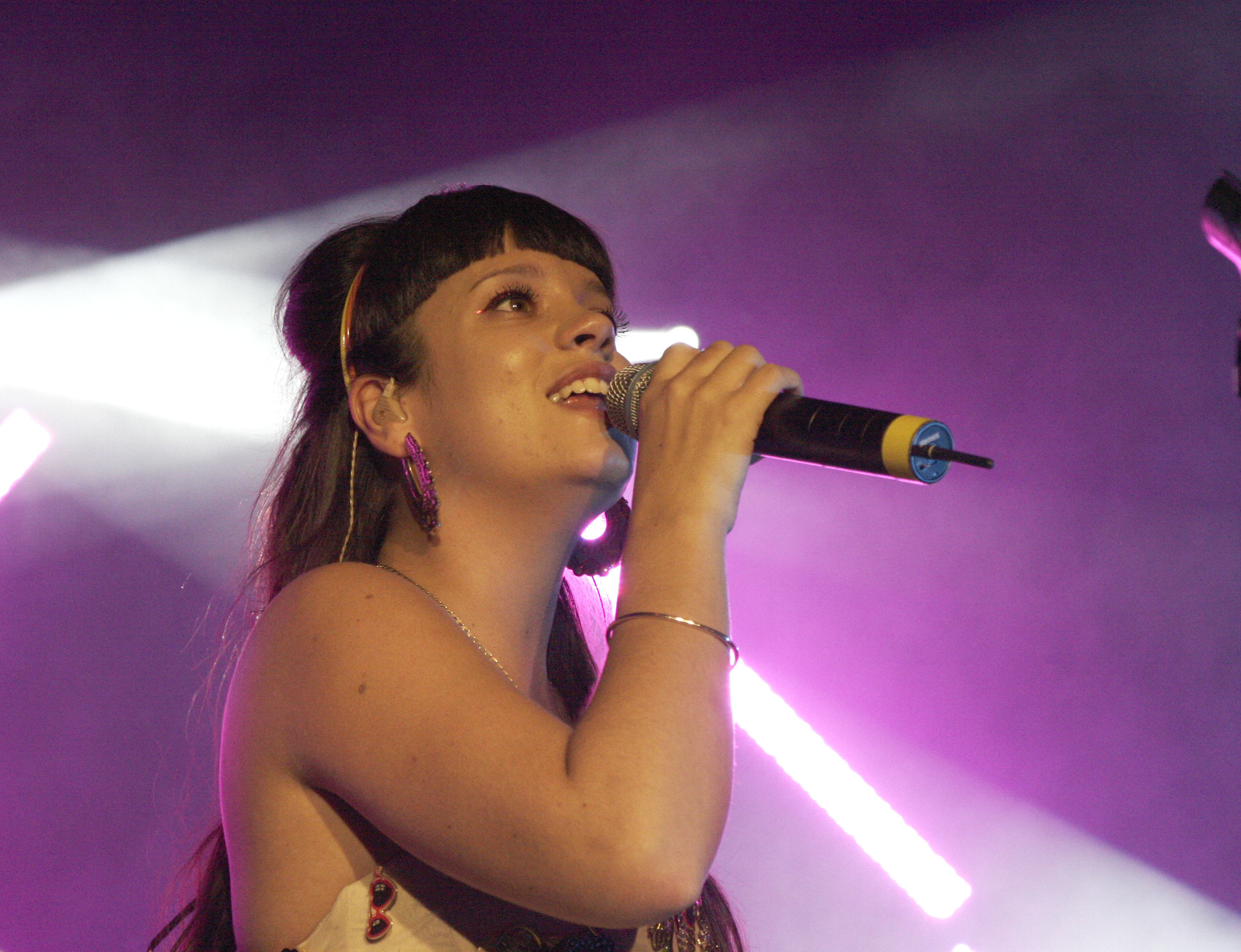 Lily Allen performing on stage with a microphone. She wears a sleeveless top and has a headband