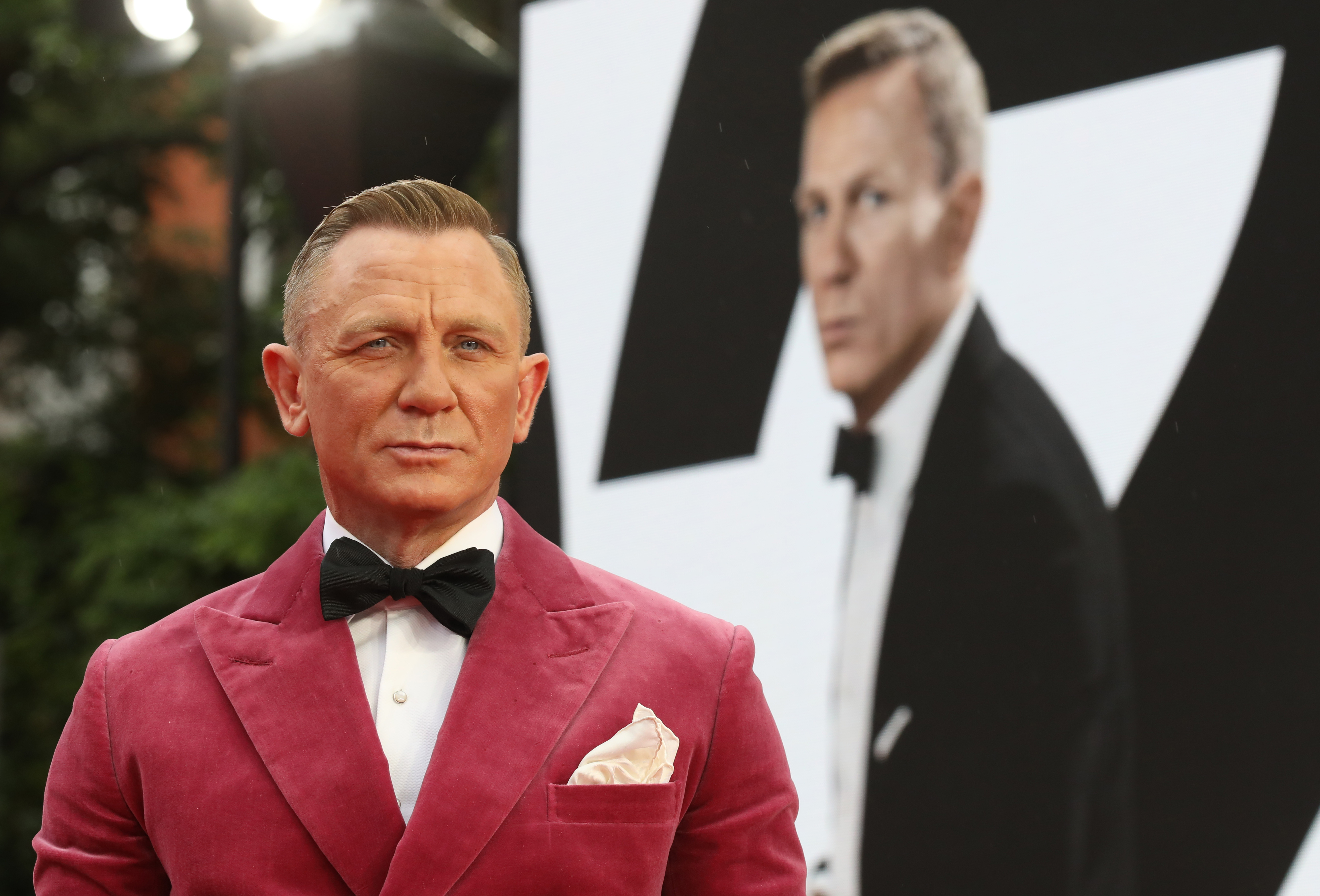 Daniel Craig in a velvet tuxedo with bow tie at a movie premiere, with a Bond poster in the background