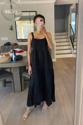 Person in a tiered black dress taking a mirror selfie with a smartphone, indoors near a staircase