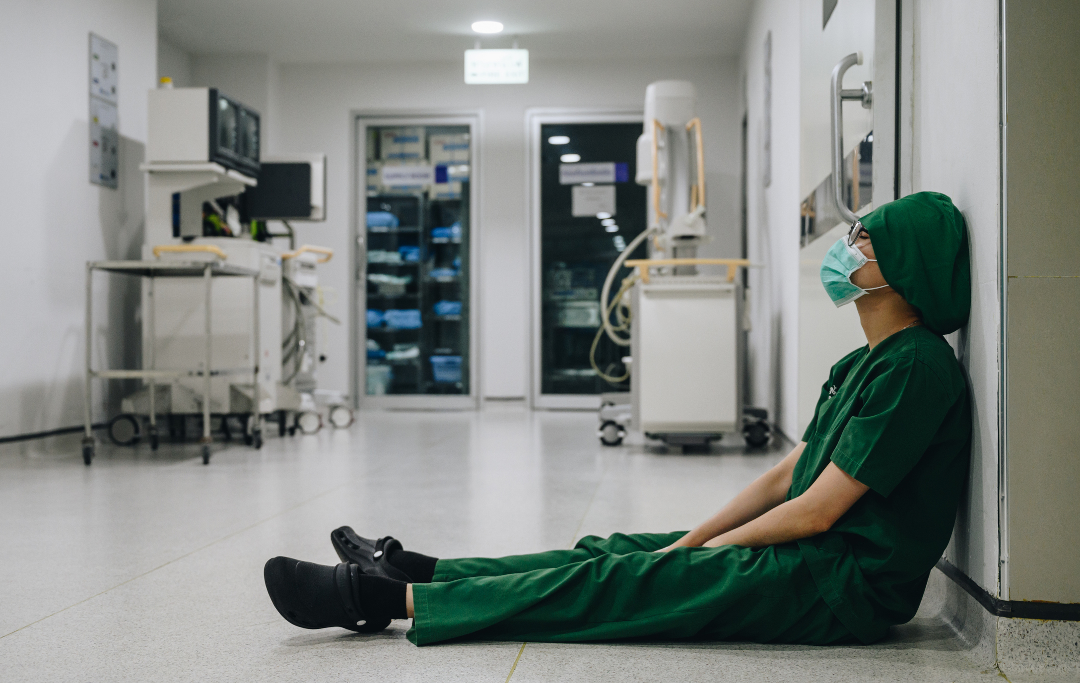 Healthcare worker in scrubs and mask sitting tiredly on the floor in a hospital corridor