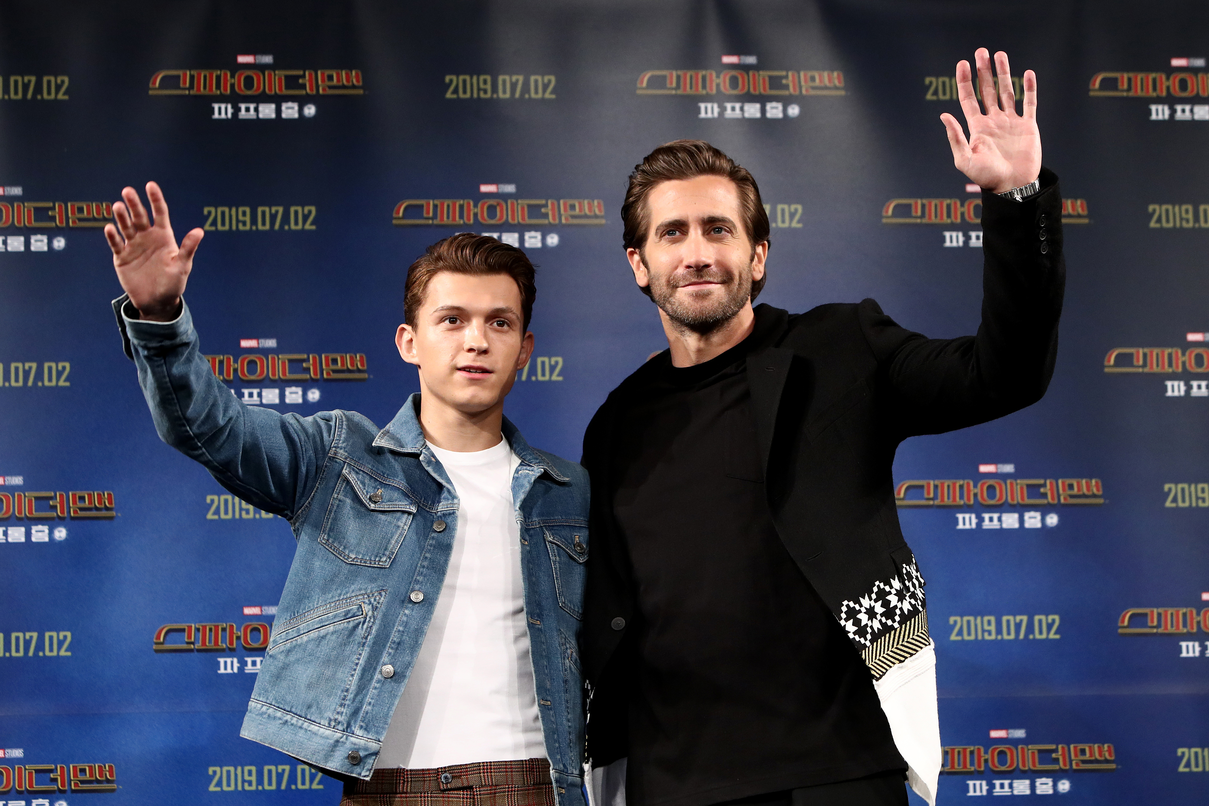 Tom Holland in denim jacket over plain shirt and Jake Gyllenhaal in a dark outfit, waving at an event