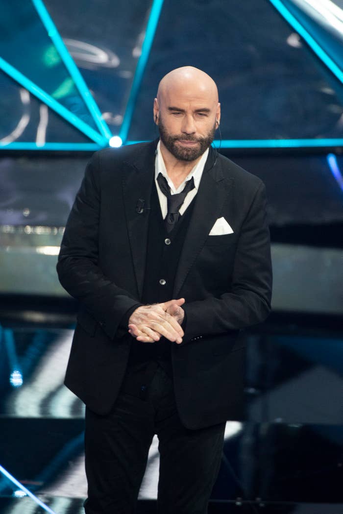 John Travolta on stage in a black suit and shirt with a white handkerchief in his pocket